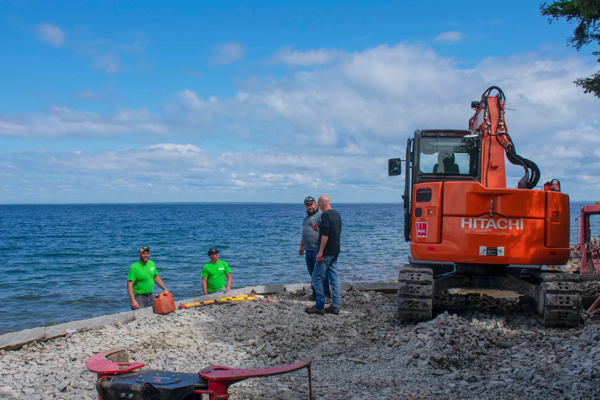Three people standing near a Hitachi excavator by the water under a blue sky with clouds.