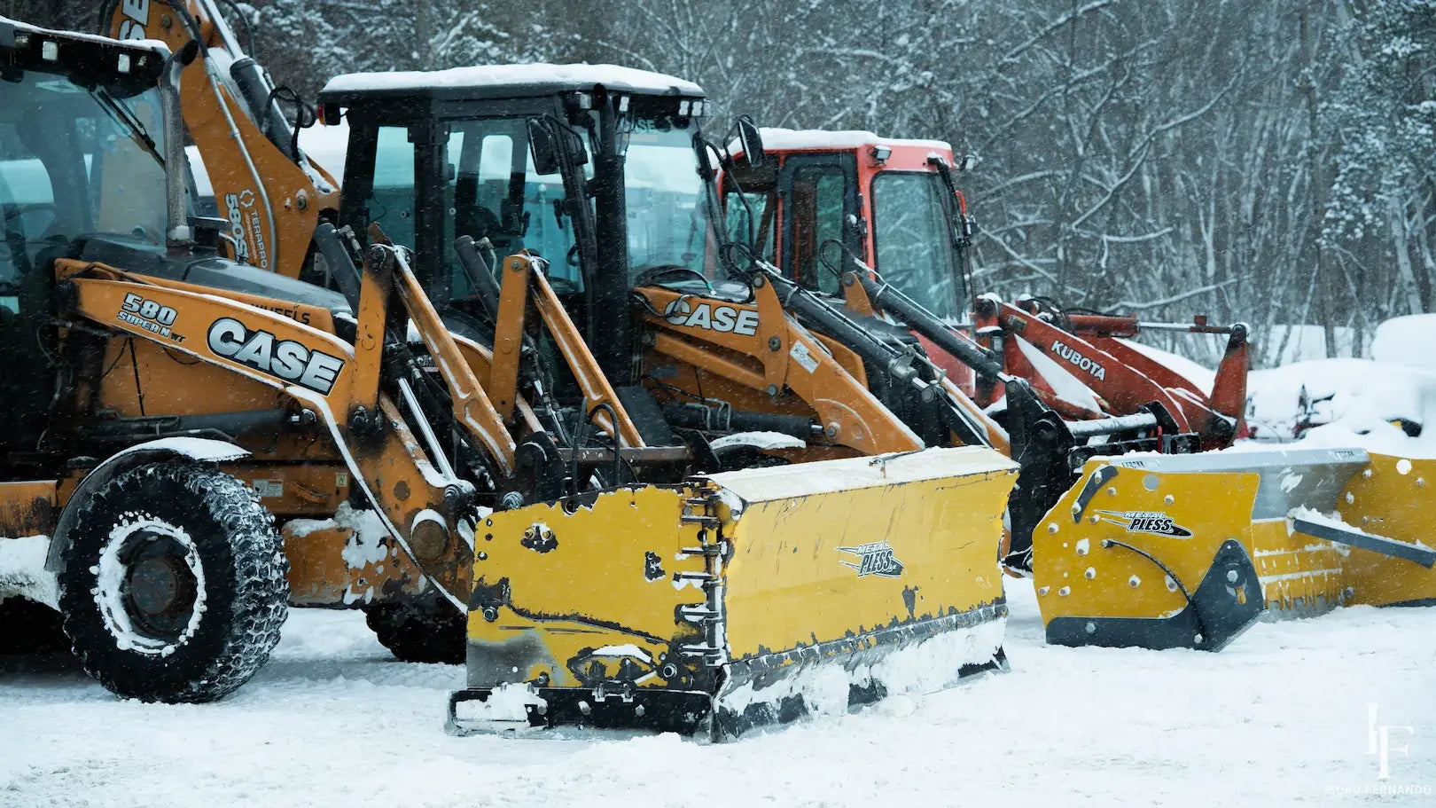 A group of heavy-duty snow removal vehicles parked in deep snow, including a prominent yellow Case 4WD snow loader with a snow blade attachment in the foreground.