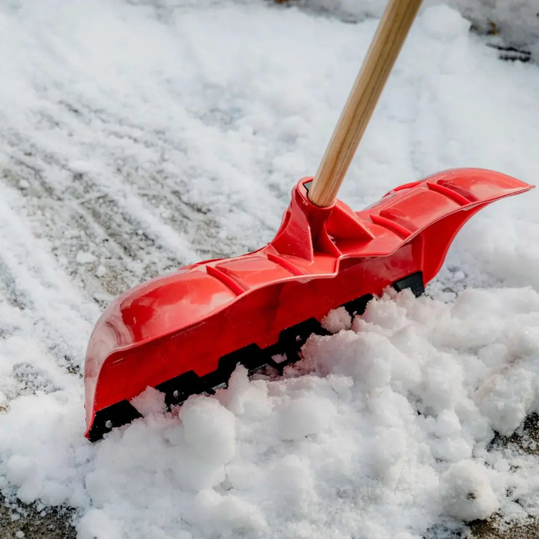 A red snowblower shovel with a wooden handle pushing snow outdoors on a paved surface.
