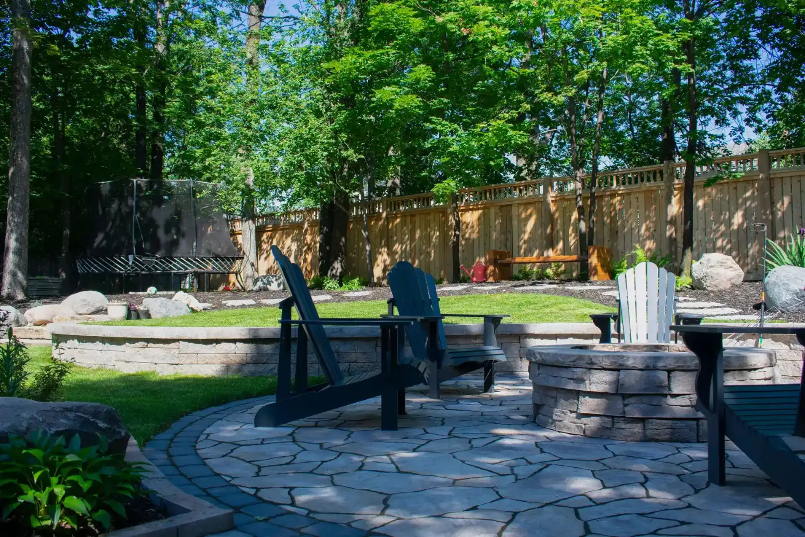 Outdoor backyard patio scene with stone pavers, two black Adirondack chairs, a stone fire pit, green grass, trees, and a wooden fence.