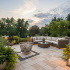 Outdoor patio with sectional sofa, stainless steel fire pit, and potted plants during sunset.